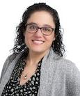 Headshot: Woman in dark top grey jacket with short brown hair and glasses smiling at camera.