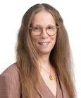 Headshot: long haired woman with glasses in med. brown top smiling at the camera.